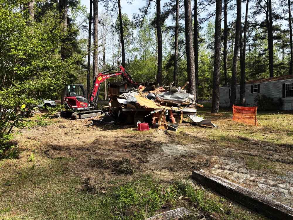 Excavator clearing a demolition debris pile at a residential site with trees.