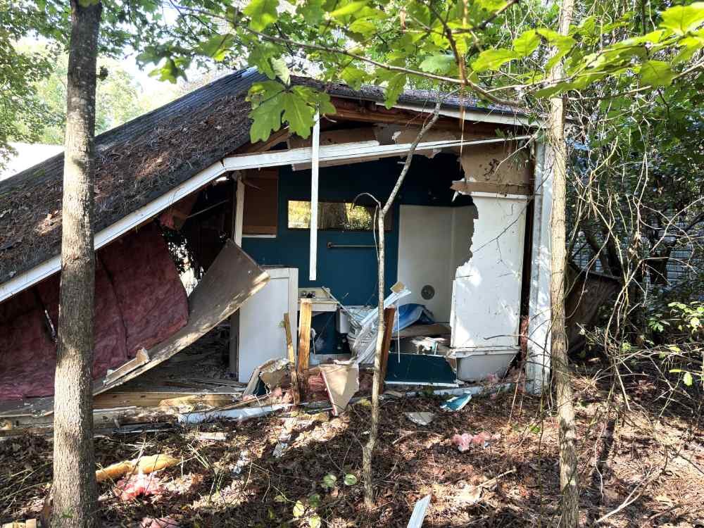 Residential structure teardown showing a collapsed roof, exposed walls, and debris in a wooded lot.