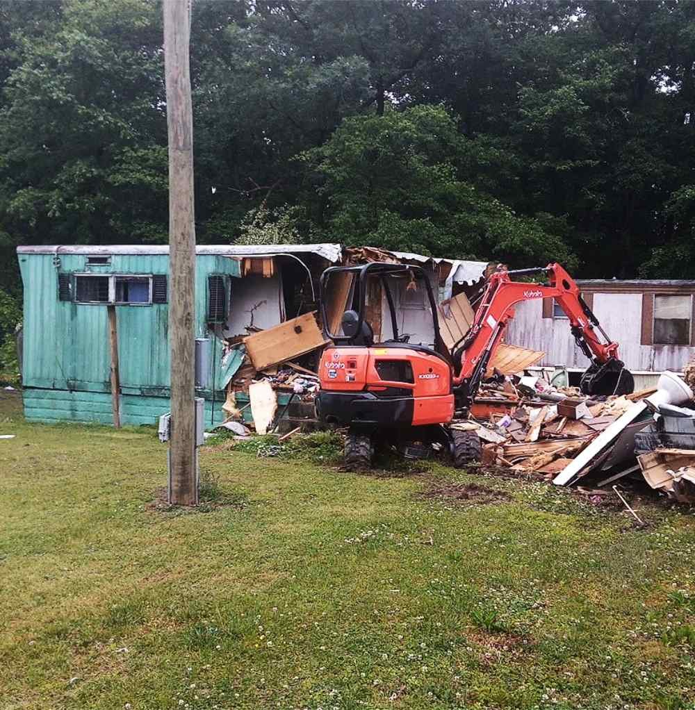Red excavator tearing down a mobile home with a debris pile during residential demolition.