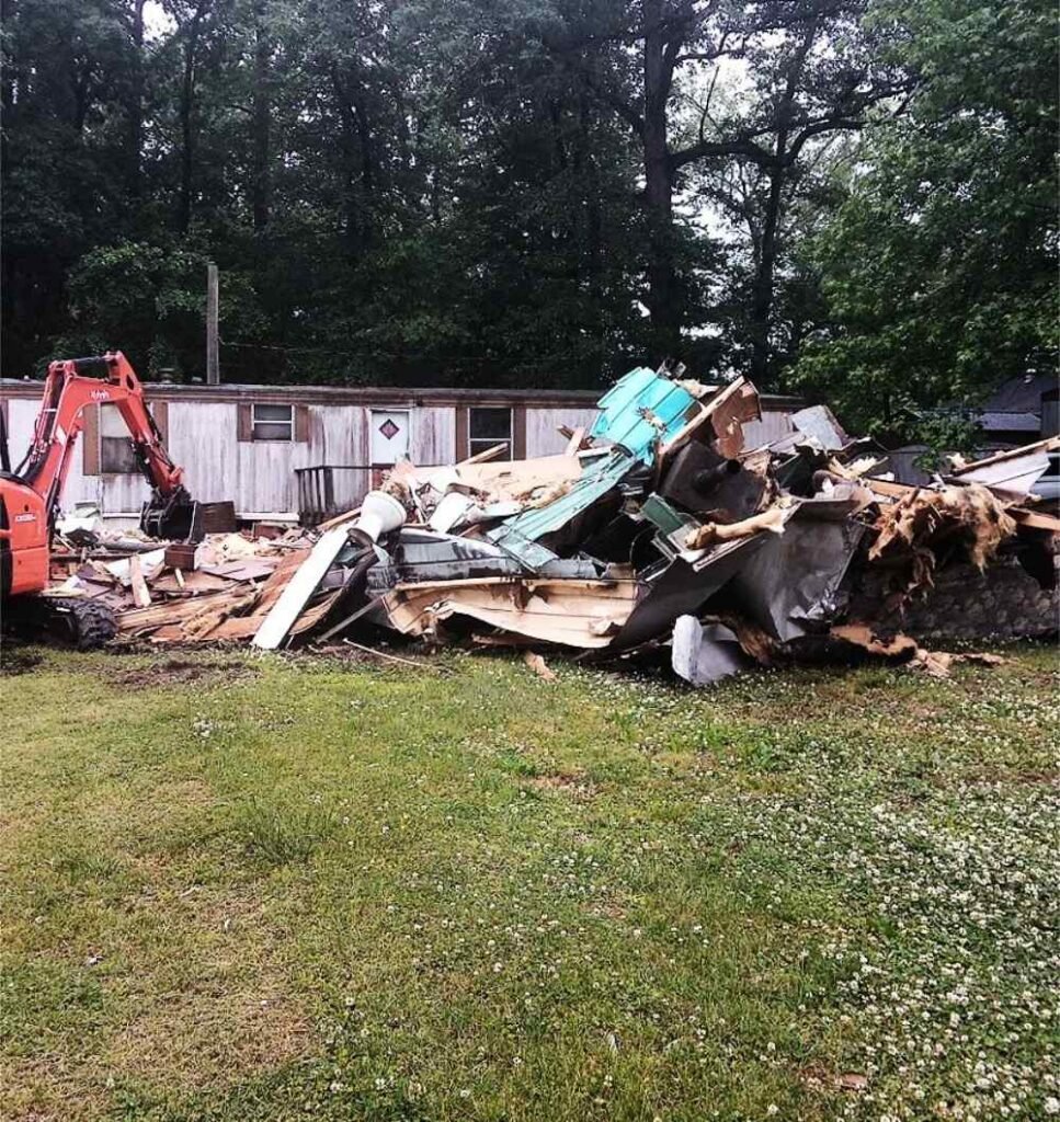 Excavator tearing down a mobile home with a large debris pile on a residential site.
