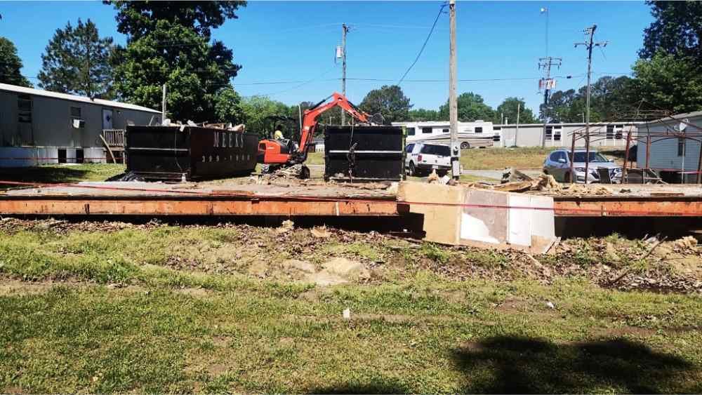 Excavator loading debris into a dumpster during a mobile home teardown and site clearing project.